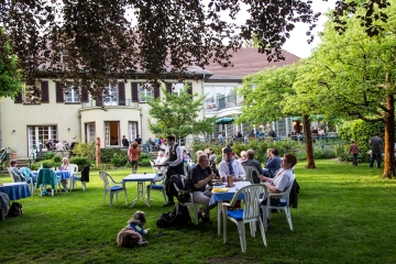 Das goldene Team 25 Jahre Hunde für Handicaps. Das Foto ist in einem gepflegten Garten aufgenommen. An diversen Tischen sitzen die Gäste unter Bäumen und haben Spaß miteinander. Die Vierbeiner haben es sich auf dem Rasen bequem gemacht.